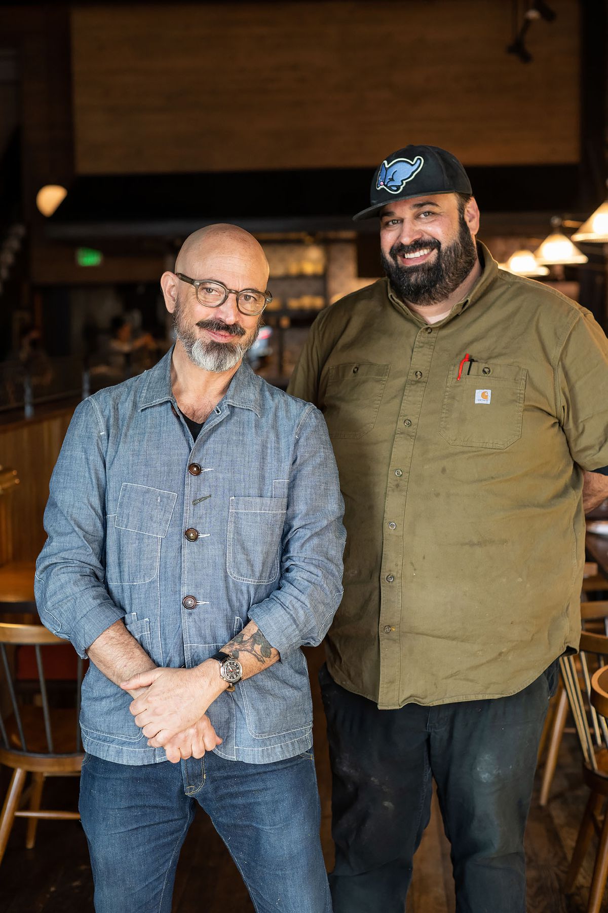 Two men wearing button up work shirt stand and smile inside a new restaurant.