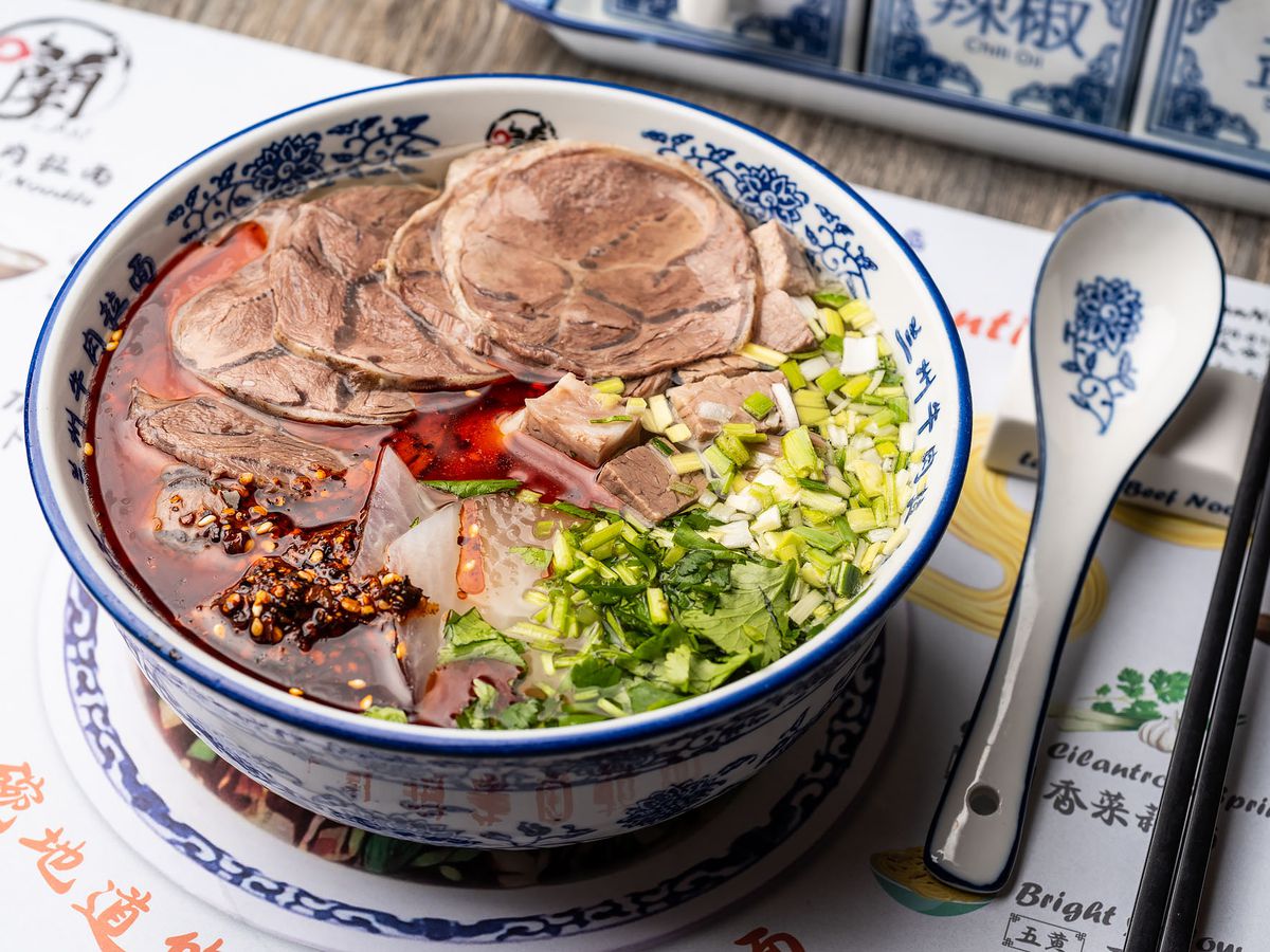 A bowl of Lanzhou beef noodle soup with cut garnishes and thinly sliced meat with a printed placemat.