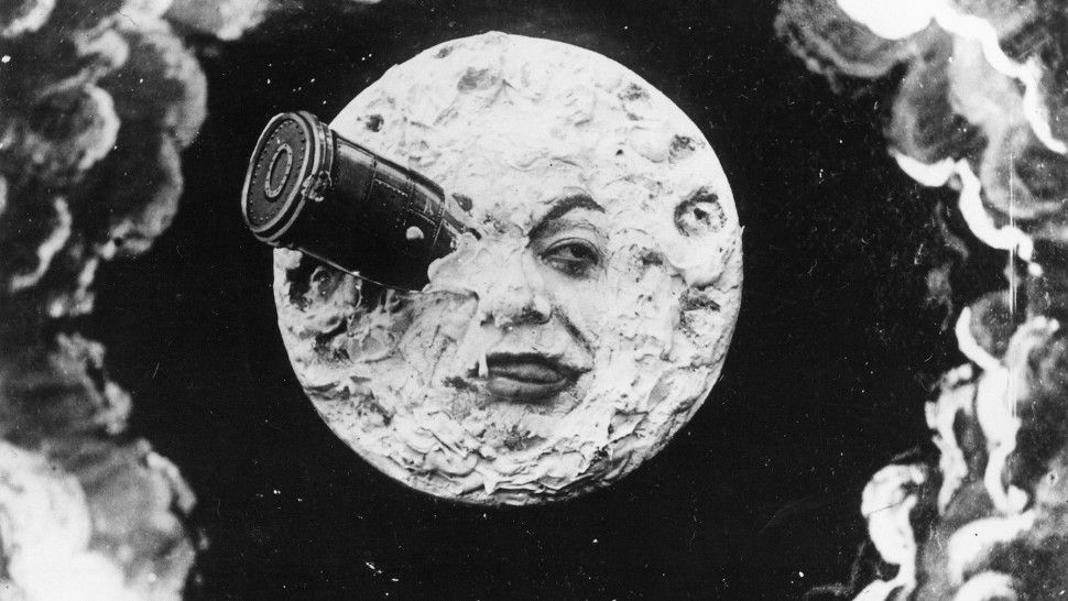 A black-and-white photo shows a man’s face looking out of a miniature moon with a telescope attached.