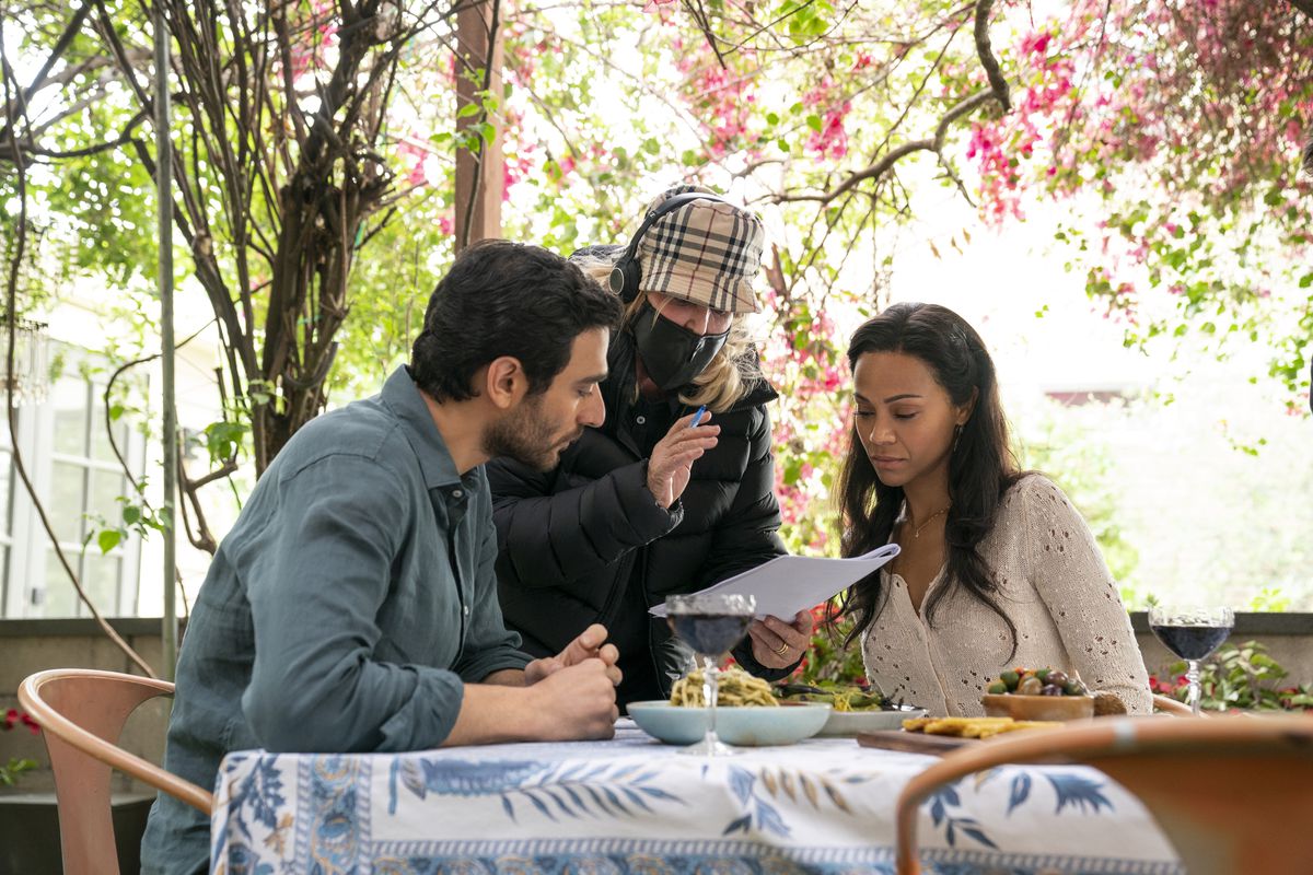 Eugenio Mastrandrea, a white man with black hair and a close-cropped beard, sits across a table from Zoe Saldana, a Black woman with long dark hair. A producer in a plaid cap and face mask stands between them.