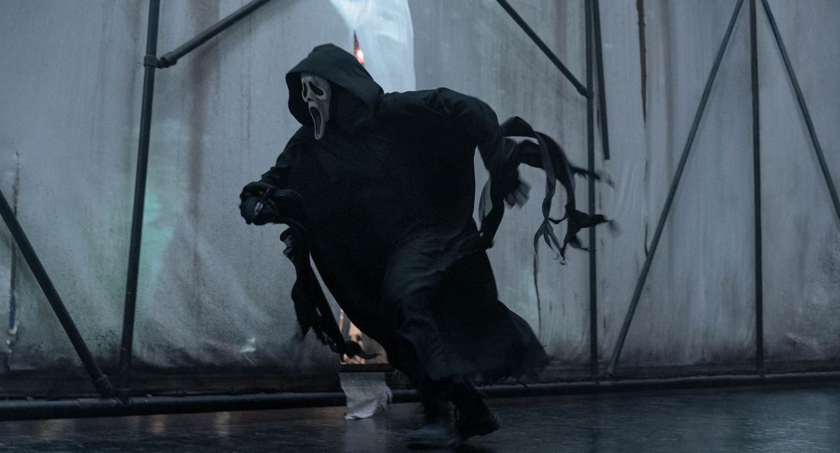 Ghostface, the black-robed, white-masked killer of the Scream franchise, runs after someone against backdrop of white scrims held up by black scaffolding in Scream VI