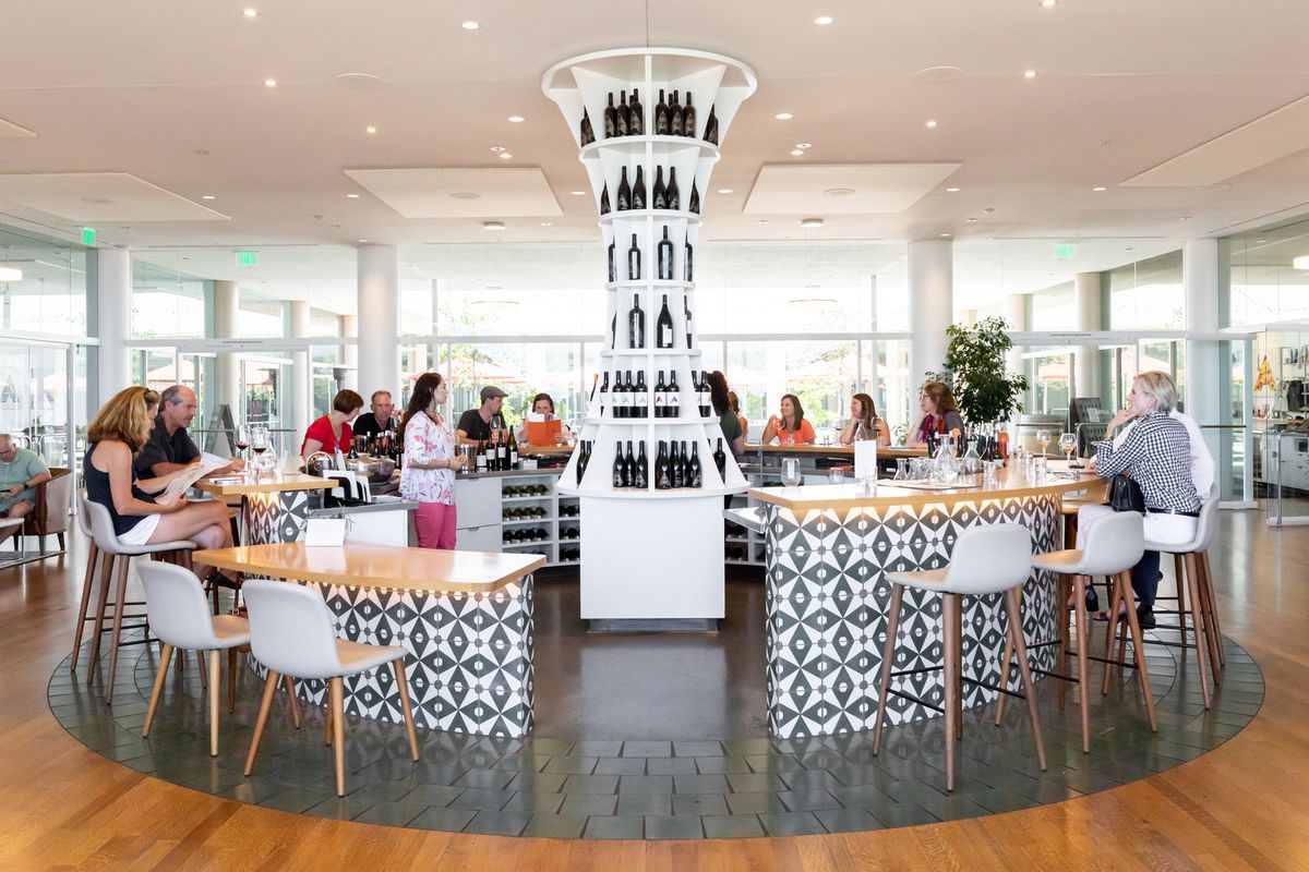 A white circular shaped bar with white chairs. The bar is decorated with white geometric shaped tiles. In the center of the bar area is a tower with shelves full of wine bottles.