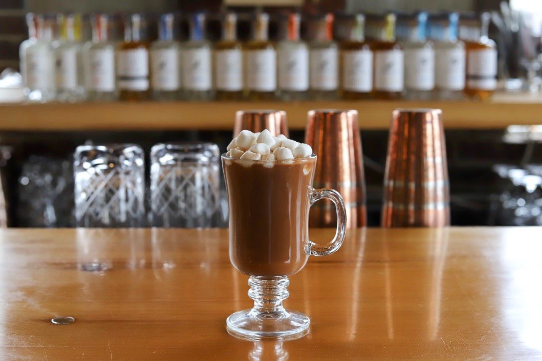 A boozy hot chocolate in a clear glass mug sits on a bar and is garnished with ample marshmallows