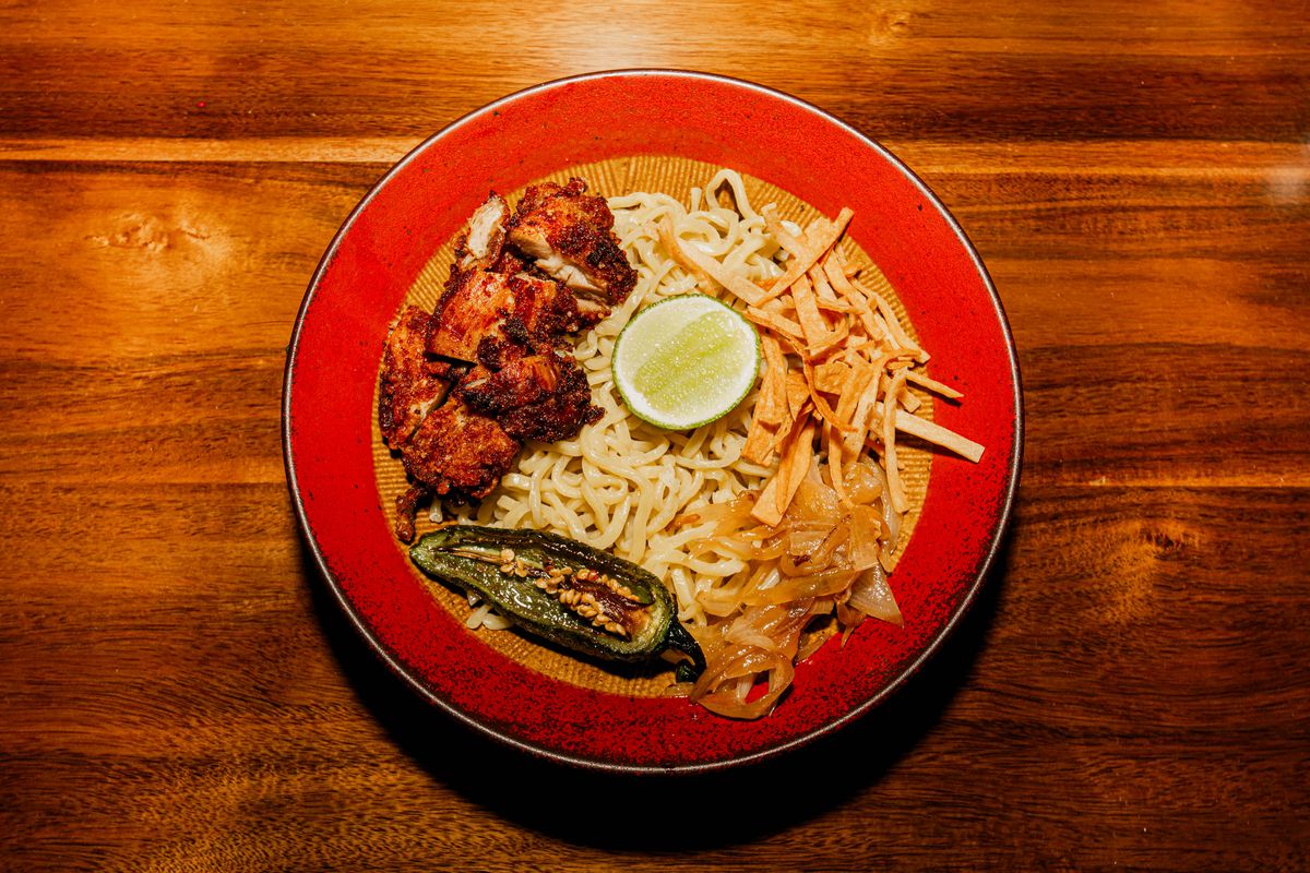 A red bowl of ramen noodles, a roasted pepper, fried chicken bites, and a halved lime. 