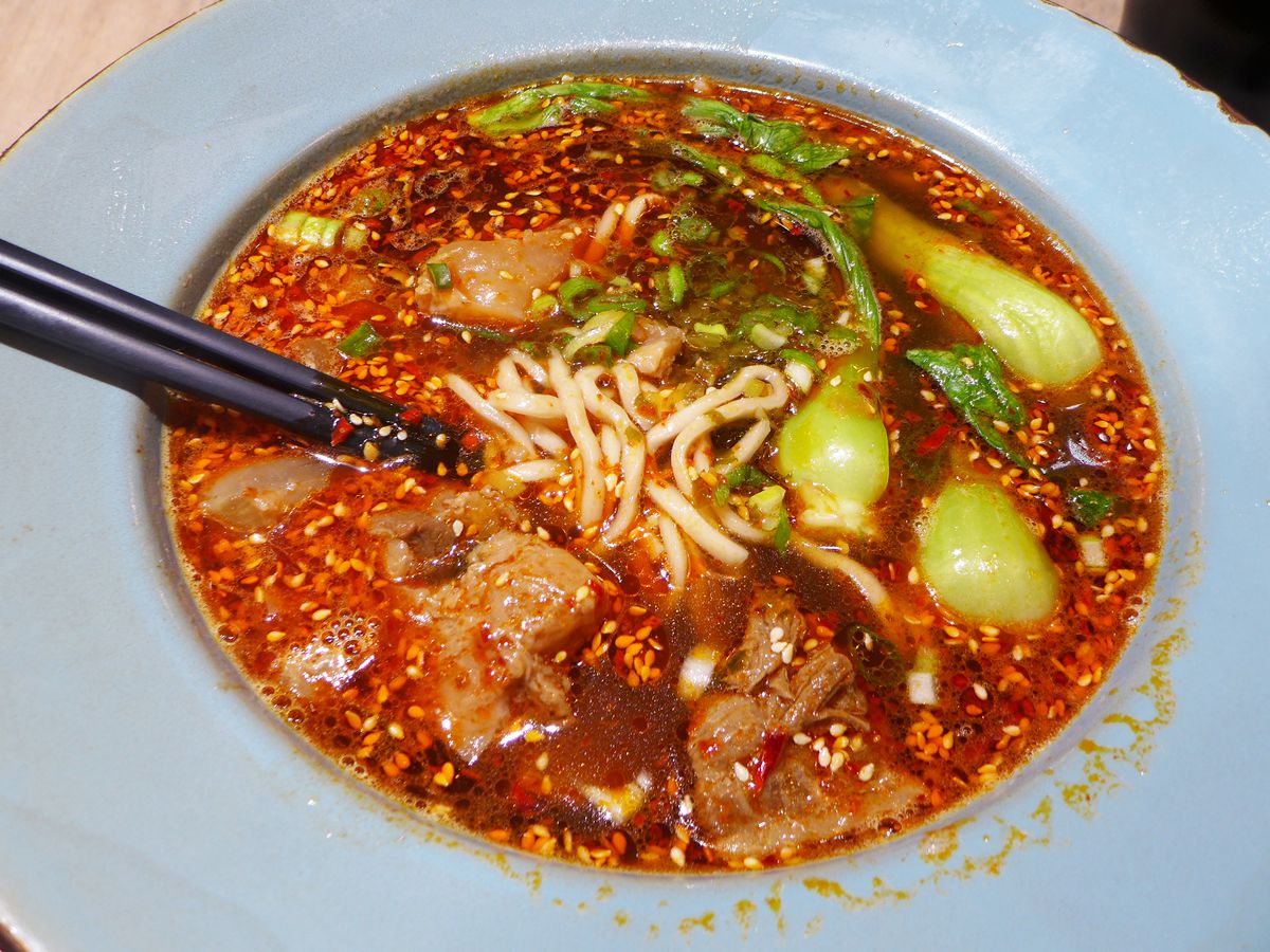 A bowl of fiery red broth and noodles in the middle being lifted up by chopsticks.