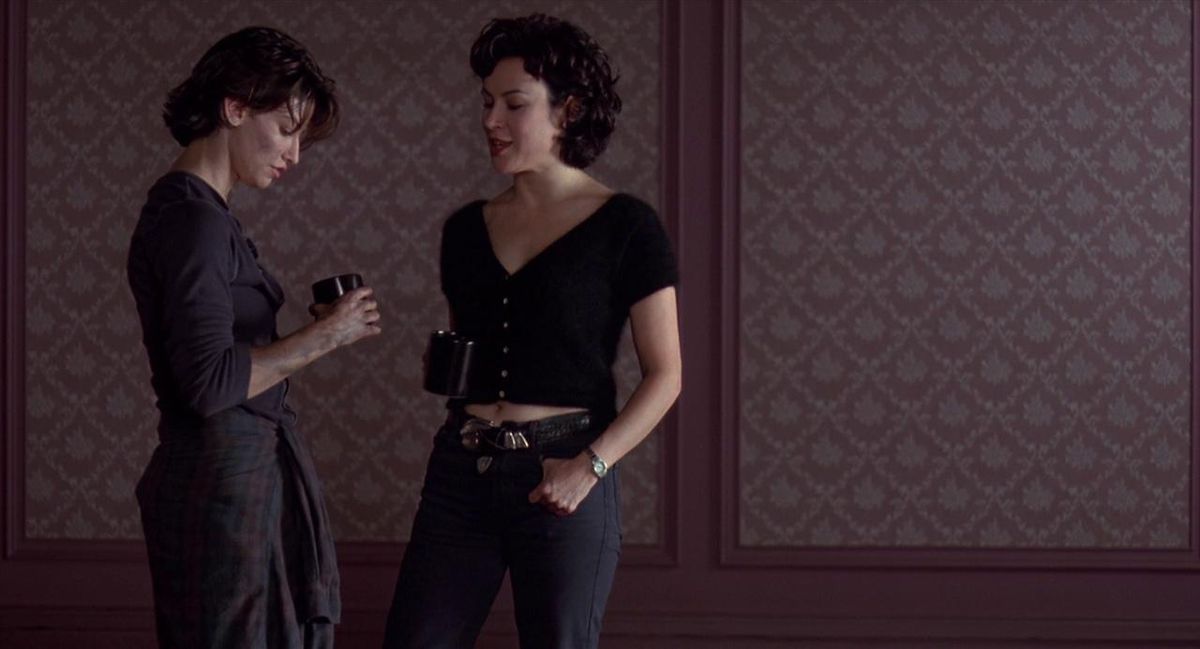 Gina Gershon and Jennifer Tilly hold coffee mugs and stand close to each other against a wallpaper backdrop in Bound.