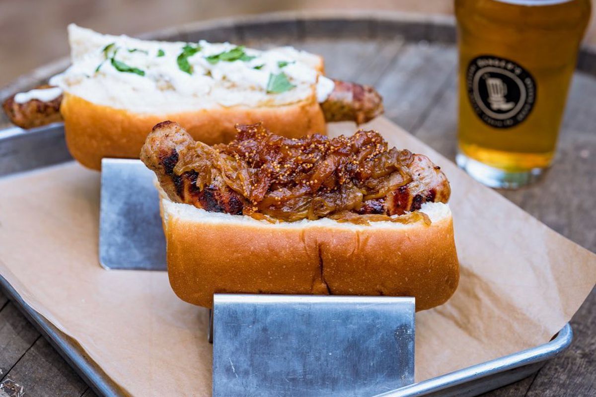 in front, a bun with barely visible sausage covered in caramelized onion, in back, bun with sausage, both in metal holders on a paper-lined tray, with a beer on the right