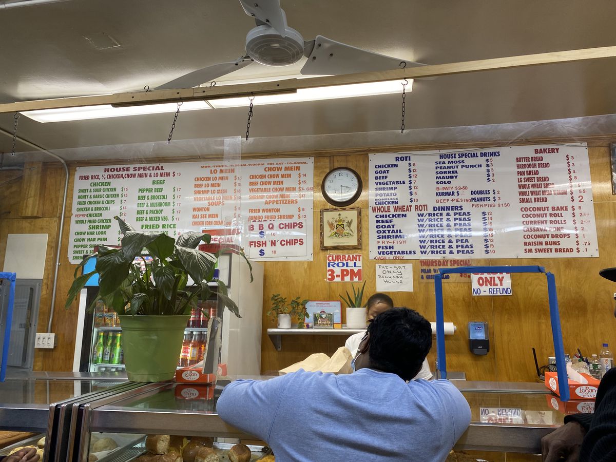 A person stands at a counter making an order from a lengthy list of specials that include chow mein, roti, and other Trinidadian-Chinese specials.