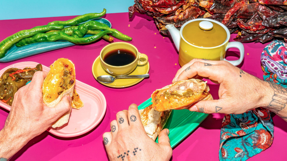 Tattooed hands rip apart a breakfast burrito next to piles of red and green chiles and a cup of coffee.