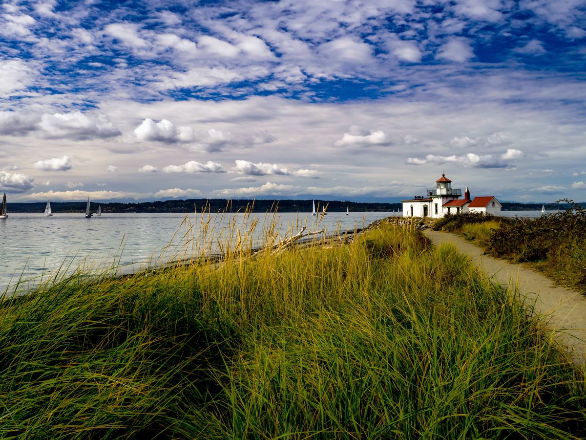 A trail surrounded by grass along a beach. In the distance is a white lighthouse with a red roof.