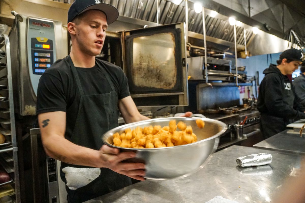A man flips tater tots in a metal bowl to season them, inside a restaurant.