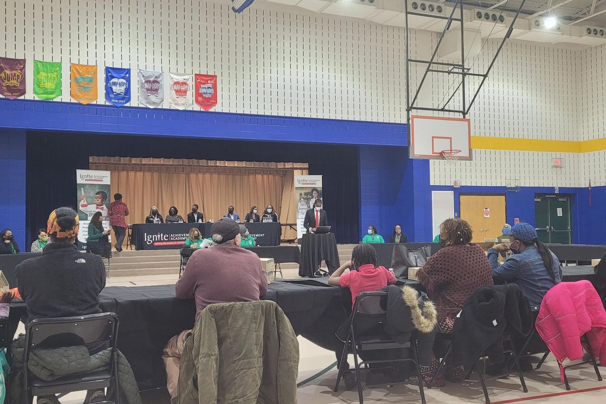 Four parents with their backs turned sit in a gym facing Ignite Achievement Academy co-founder Shy-Quon Ely as he gives a speech on the status of the school, with board members on a raised stage behind him.