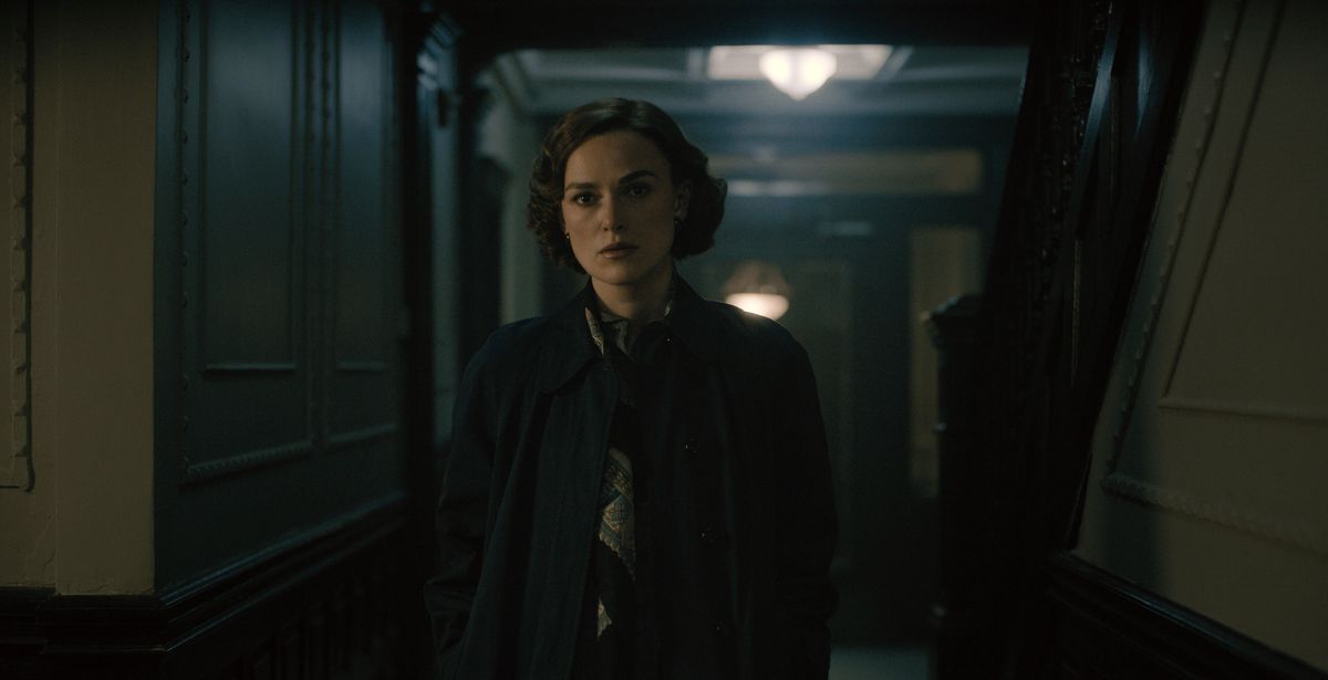 A woman (Keira Knightley) in a dark blue coat stands in in a darkly lit hallway