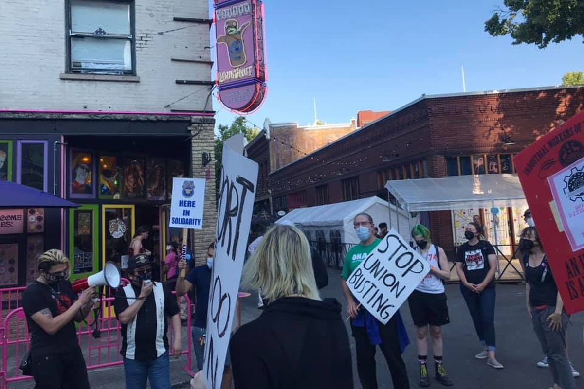 Around 20 employees stand outside Voodoo Doughnut’s location in Old Town Portland holding signs that read “Stop Union Busting.”