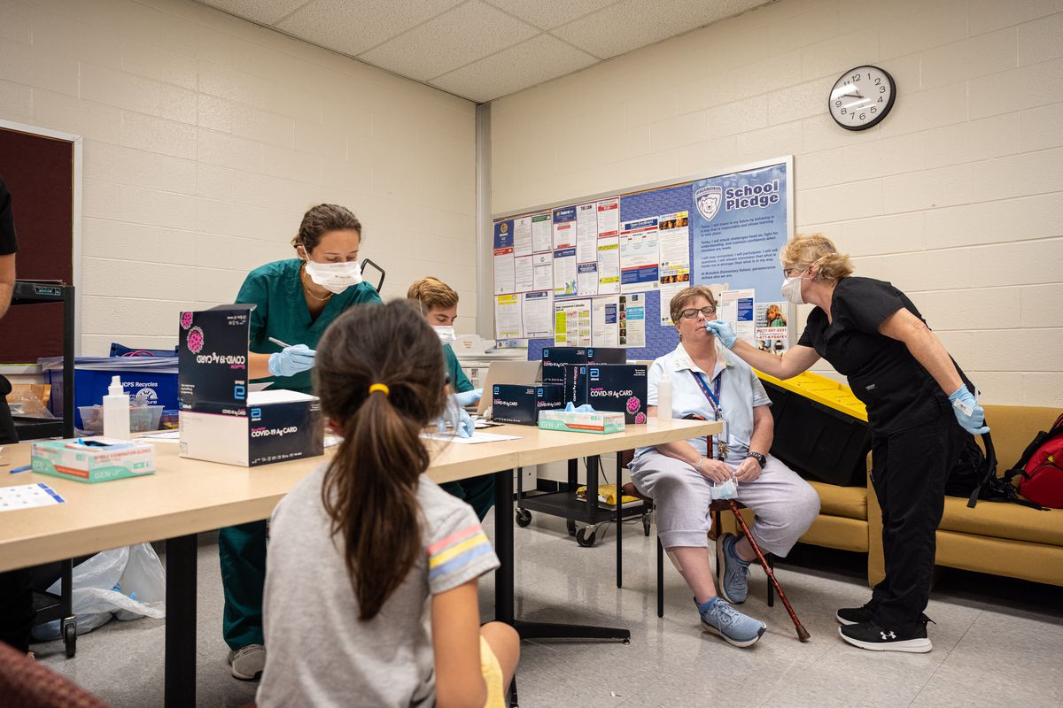 Health care professionals conduct COVID testing in an office with white brick walls.