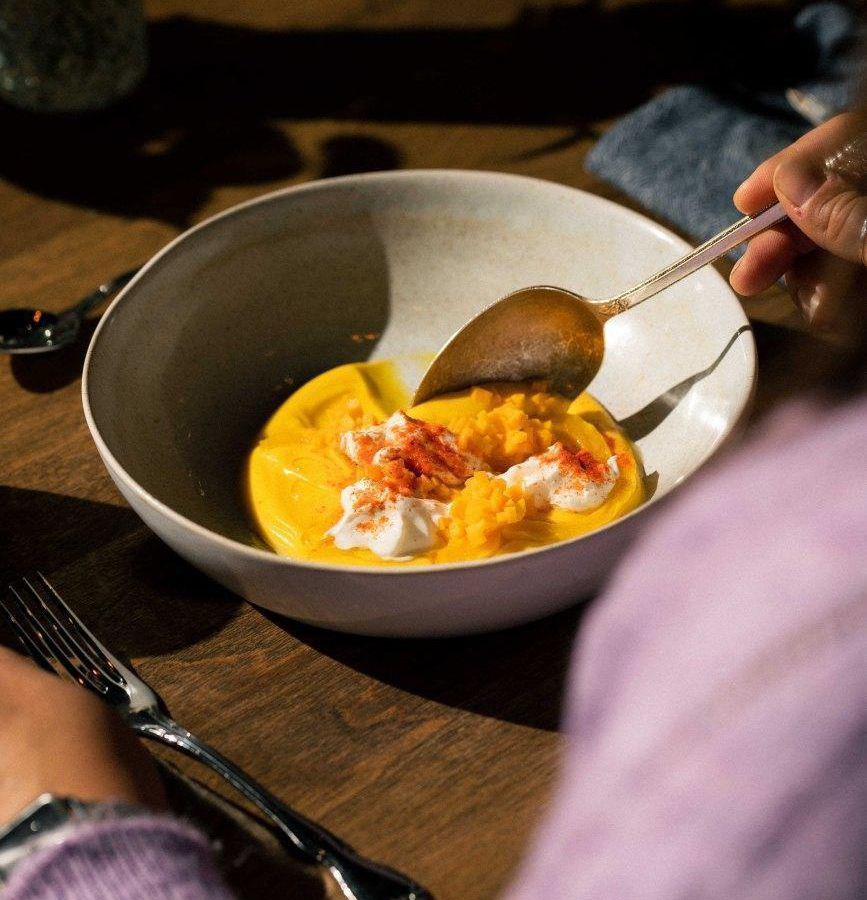 A diner digs a spoon into a bowl of thick yellow sauce topped with creamy cheese and other fixings. 