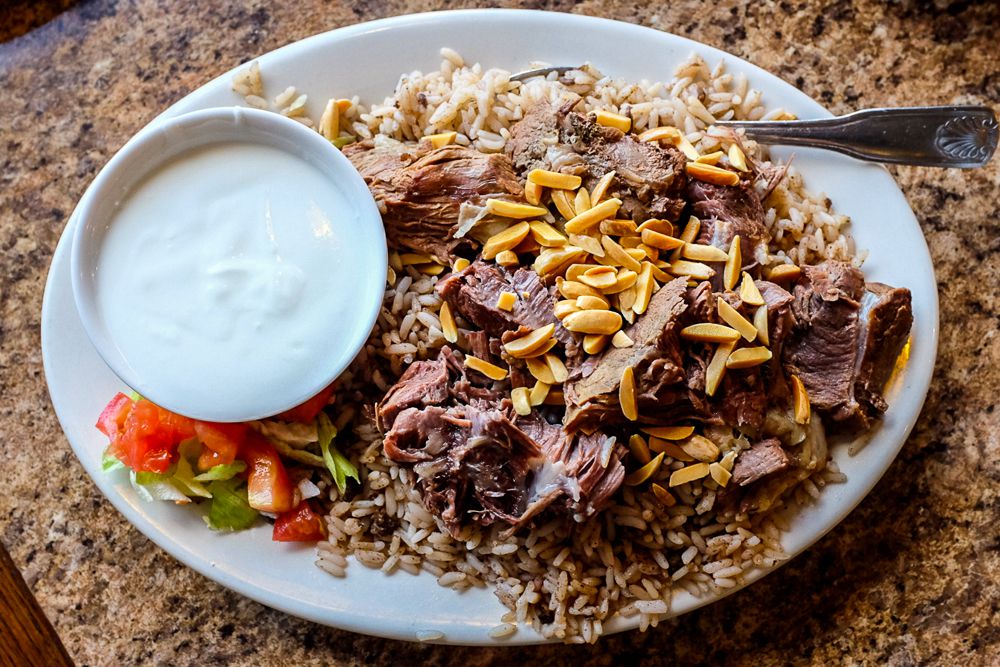 A large plate of rice with stuffed lamb pulled into pieces on top and sprinkled with slivered almonds. A large dish of creamy white sauce sits on the plate next to some cubed tomatoes. 