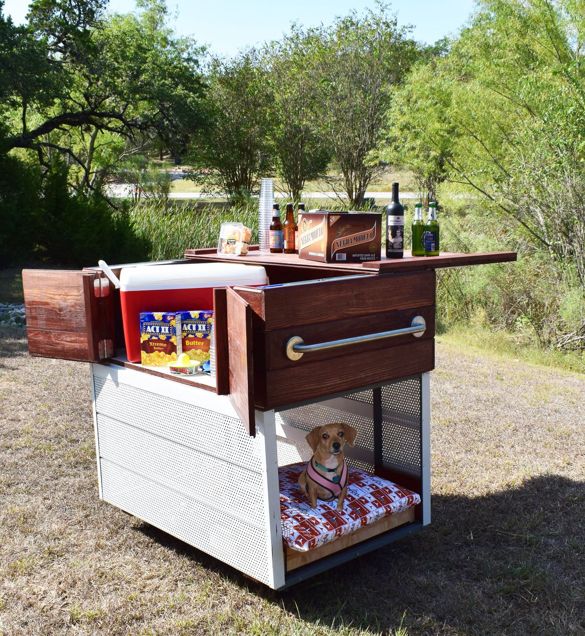 A two-tiered rolling cart. The bottom has perforated metal walls painted white, with a small dog on a pillow inside. The top is wood and designed to serve as a bar or serving area. There are popcorn boxes and beer bottles on top.