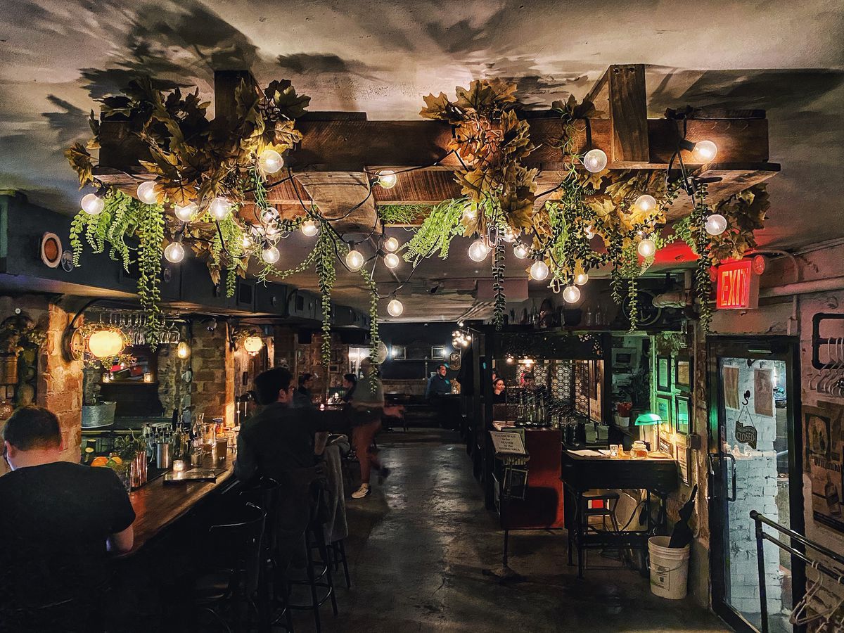 The interior of a dark restaurant with black floors, plants and light hanging from the ceiling, and rows of wooden tables and chairs
