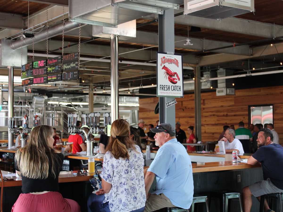An industrial style bar interior with patrons seated around a central bar with many taps, metal ceiling and lights, and an illustrated sign reading “Narragansett Fresh Catch” with an image of a lobster