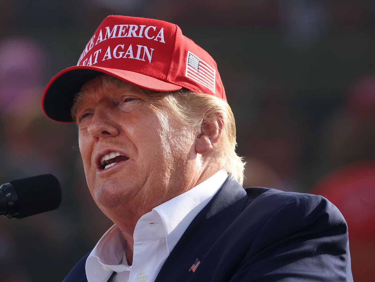 Donald Trump speaking into a microphone while wearing a “Make America Great Again” hat.