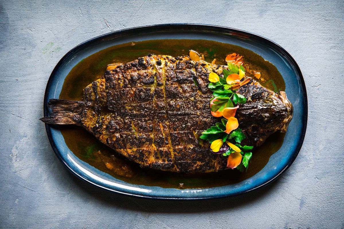 A whole blackened fish sits on a blue plate at Kann, with a lei of herbs and edible flowers.