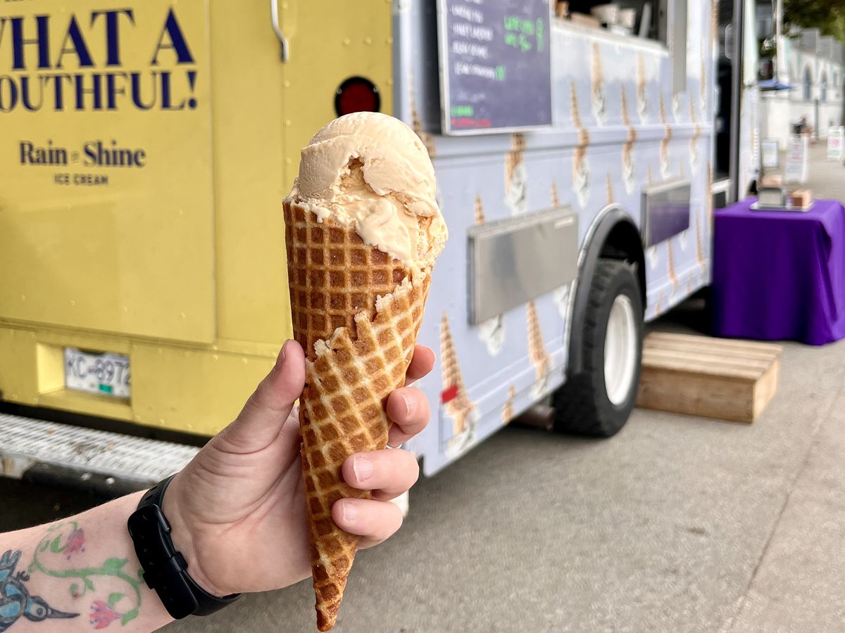 A hand holds an ice cream cone in front of a food truck.