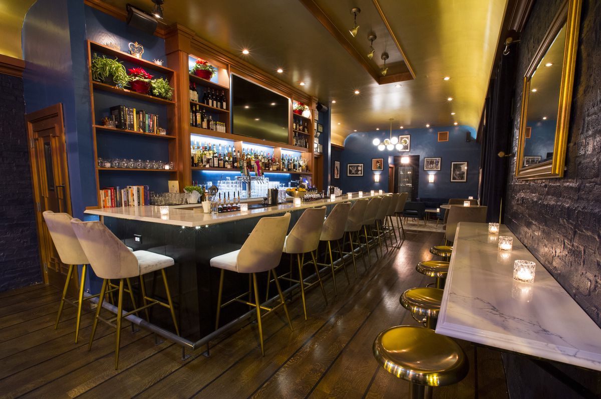 A blue bar space with a long white bar and brass stools at small tables along the walls.
