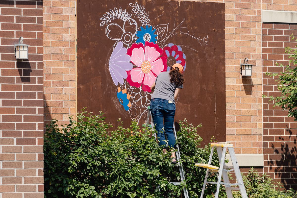 A woman paints a floral mural on the side of a brick building
