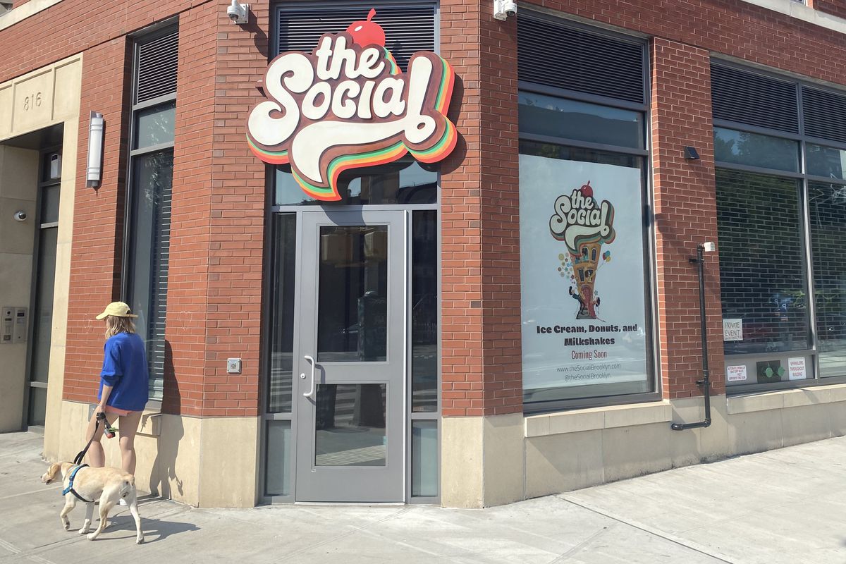 A person wearing a blue sweater and a yellow hat walks a dog past an unopened storefront with a colorful sign that reads “the Social”