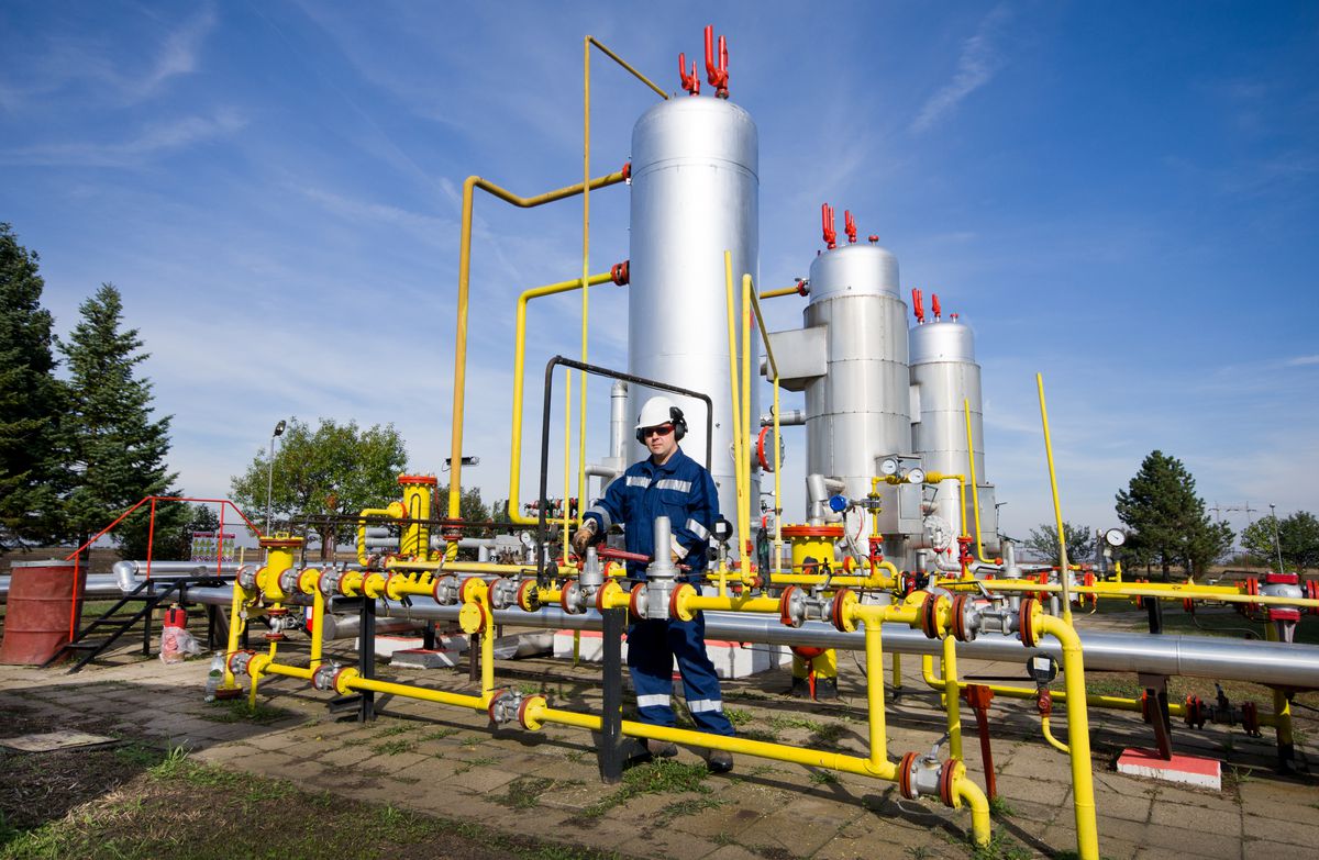 A man in work clothes stands amid a curving mass of pipes at a natural gas plant. 