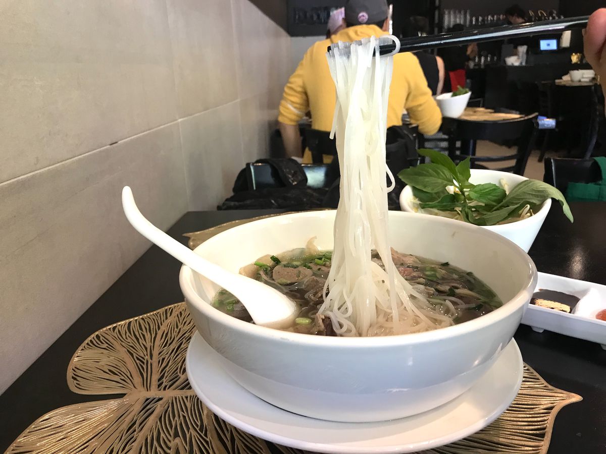 A white bowl with a light brown broth with thin rice noodles, some lifted with a pair of chopsticks.