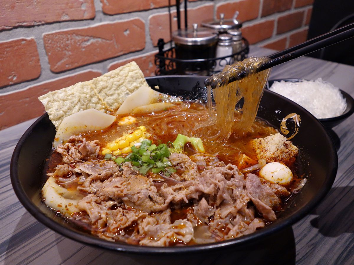 A bowl of vermicelli noodles, sliced beef, and other meats and vegetables in chili-red broth in a black bowl.