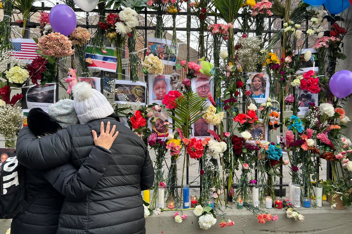 Two people in winter clothing embrace next to a memorial for the victims of a fatal fire in a Bronx apartment. The memorial is adorned with flowers, candles, and photographs of the victims.