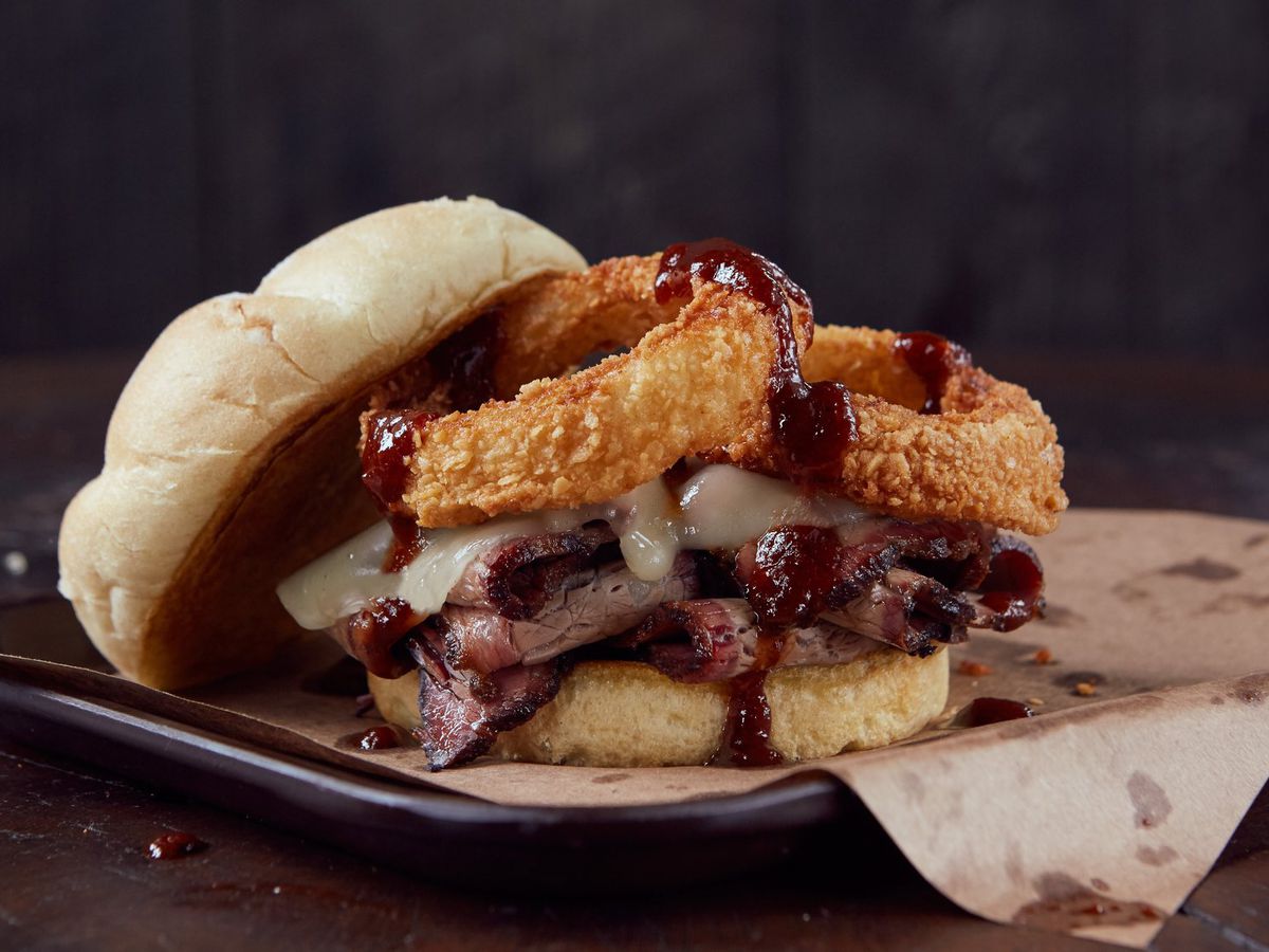 A hulking sandwich, its top bun pushed aside to reveal slices of brisket topped with melted cheese, onion rings, and drizzles of sauce, sitting on a grease-stained parchment paper-lined metal tray