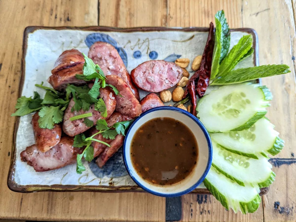 Overhead view of a Thai-style sausage, sliced and served with cucumbers, mint, peanuts, chiles, and a dipping sauce.
