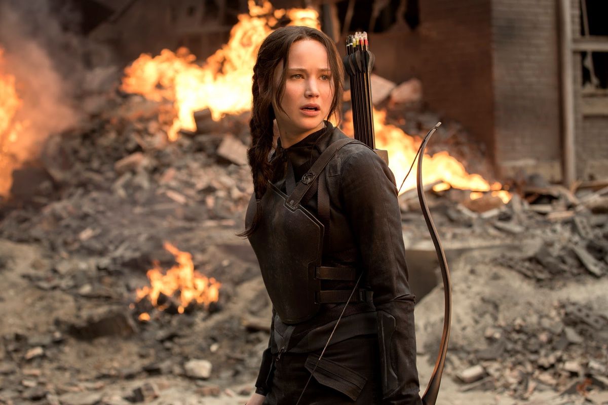 The Hunger Games - Katniss Everdeen standing in rubble with a bow slung over her shoulder and fire behind her