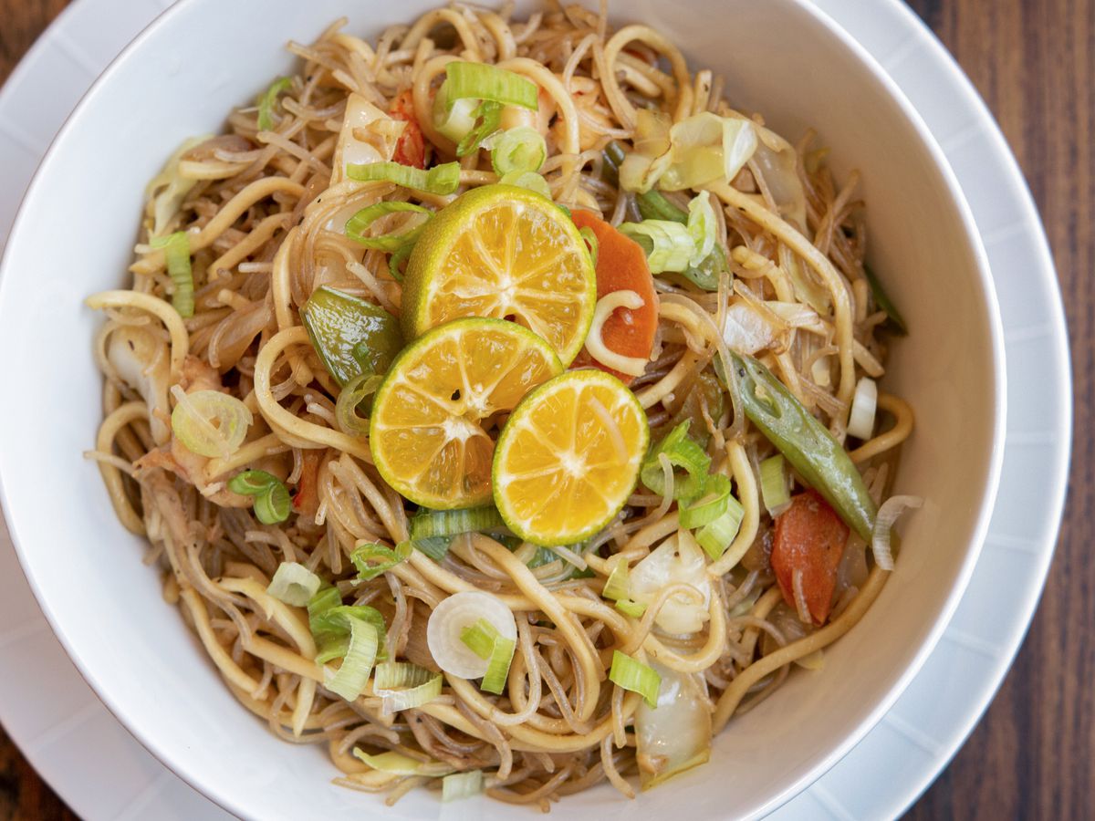 A bowl of pancit or Asian vermicelli sauteed in a wok with assorted vegetables, garnished with green onions and three slices of lemon