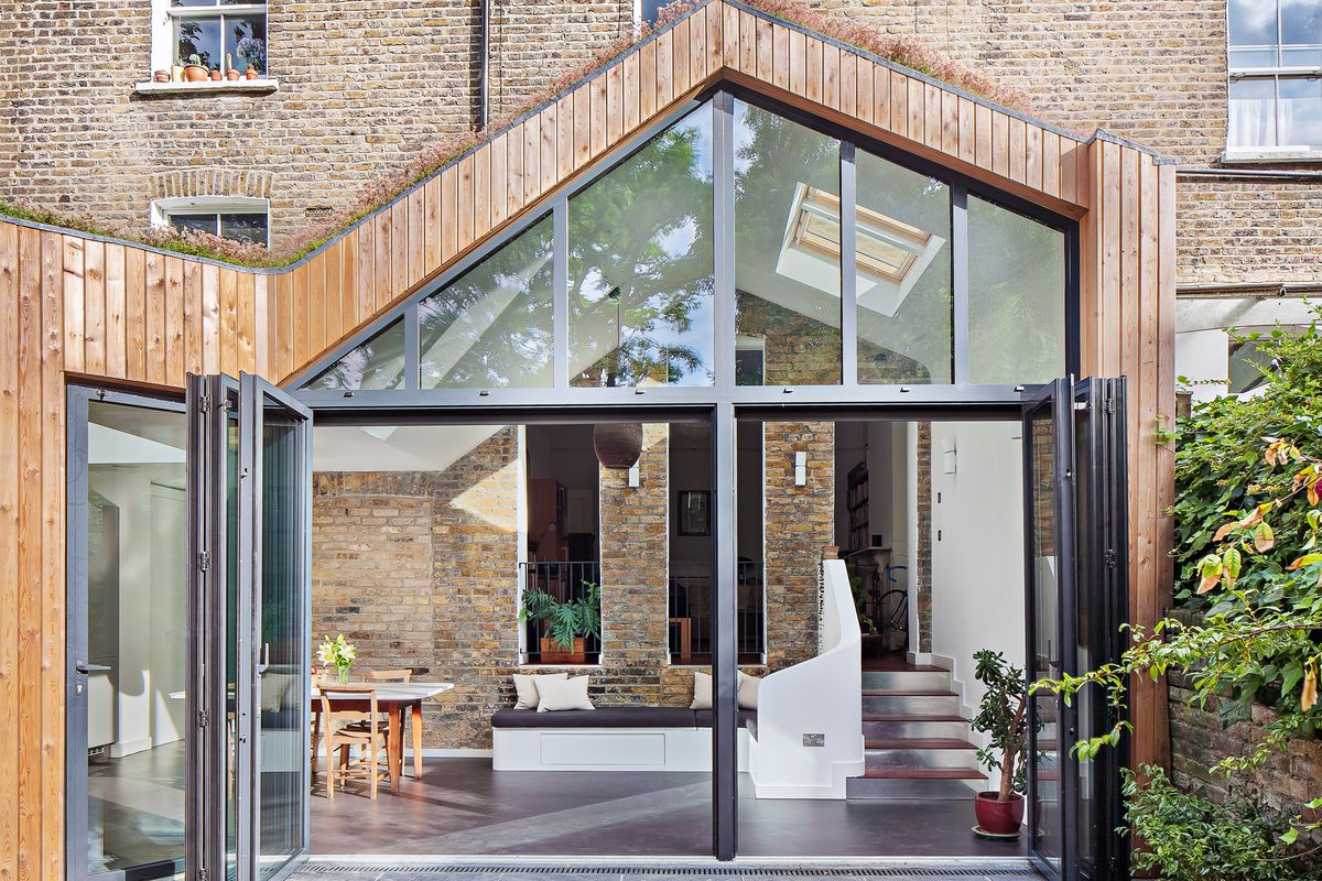 View of an extension with sloping, asymmetric roof with a wall of windows and accordion glass doors opening onto a bright kitchen and dining space. 