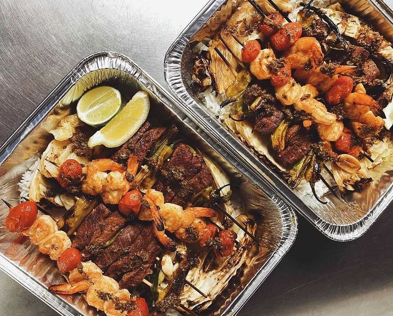 Two rectangular aluminum tins are full of steak and shrimp skewers over a bed of braised cabbage