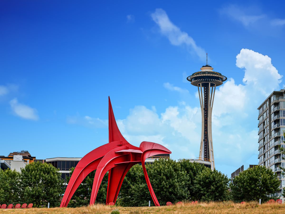 An abstract red sculpture with four curved legs and a point on top on a lawn in front of low shrubbery. An office building and a tall, pointy building (the Space Needle) are in the background. The weather is sunny with a few clouds.