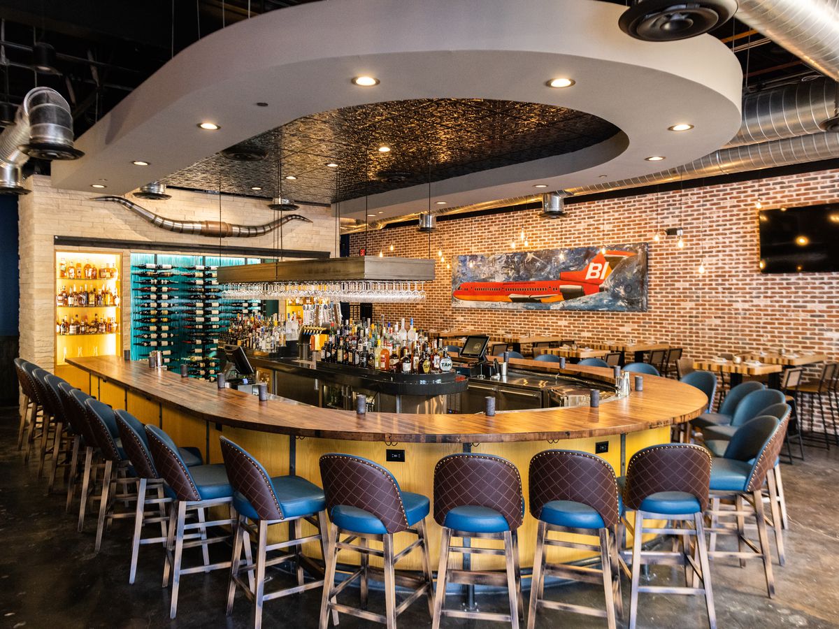The U-shaped bar inside District in Addison. It’s lined with blue and brown barstools and the bartop is made of blonde wood. A U-shaped ceiling feature mirrors the bar.