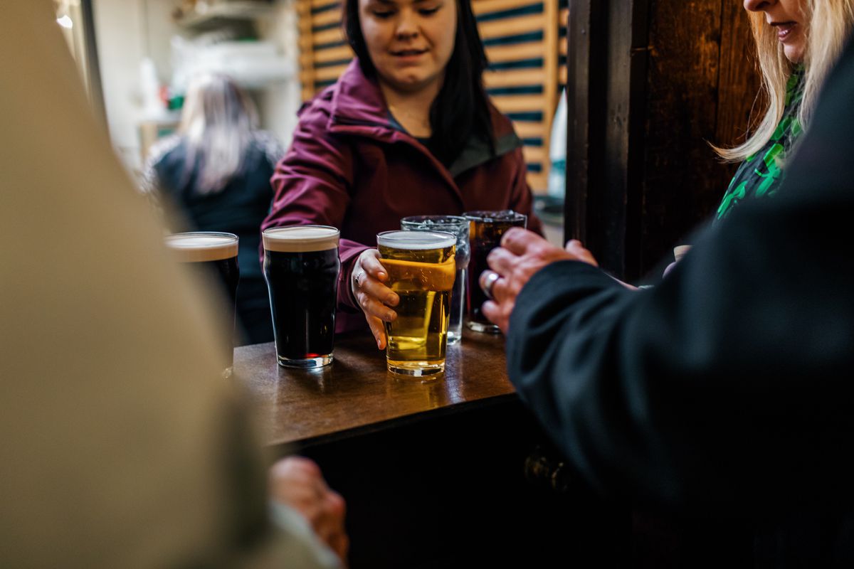 A woman in a sweatshirt passes pints across the bar to waiting customers. 