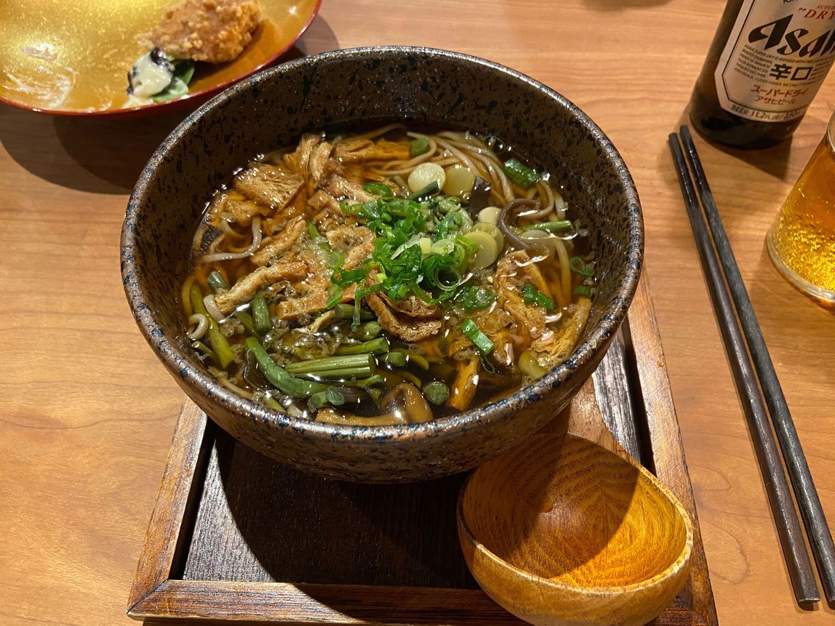 A bowl of noodles with soba, scallion, and fried tofu shreds sits atop a wooden square base with a wooden spoon.