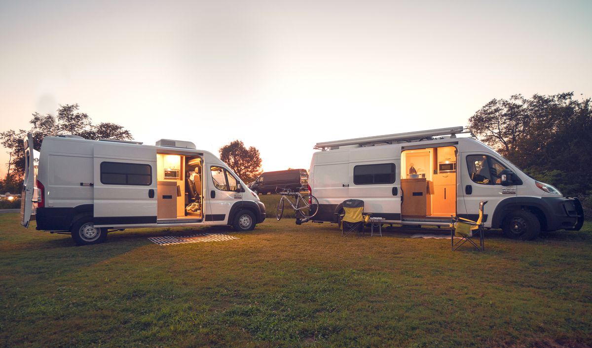 Two white camper vans sit in a grassy area with trees in the background. It’s sunset, so the van doors are open and you can see the light from within.