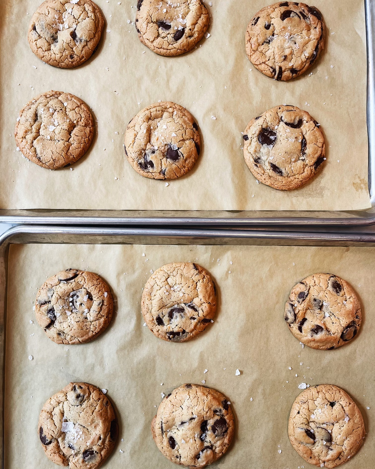 A dozen chocolate chip cookies on 2 baking sheets with parchment paper underneath and flakes of salt scattered across.
