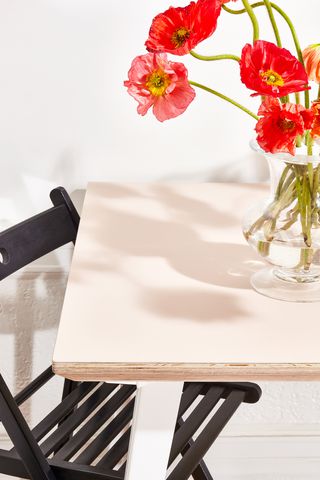 een lichtroze tafel met witte poten, een zwarte klapstoel en rode klaprozen in een heldere glazen vaas.