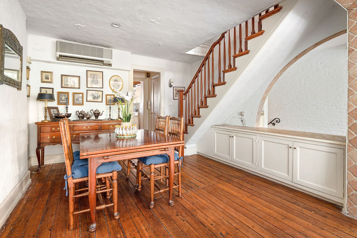 A dining area with hardwood floors, a dining table, four chairs, white walls, and a wooden staircase in the back.