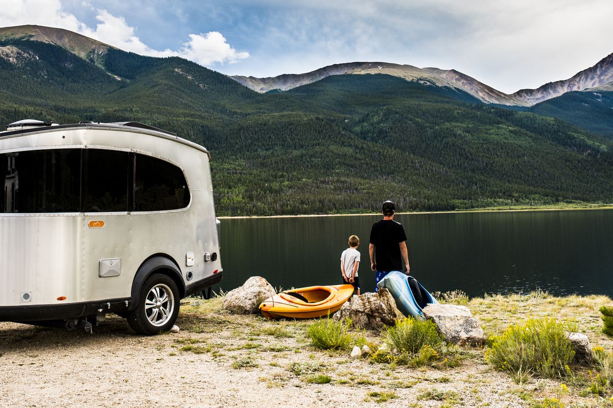 A compact silver and black-windowed trailer sits unhitched by a lake and mountain scene. 