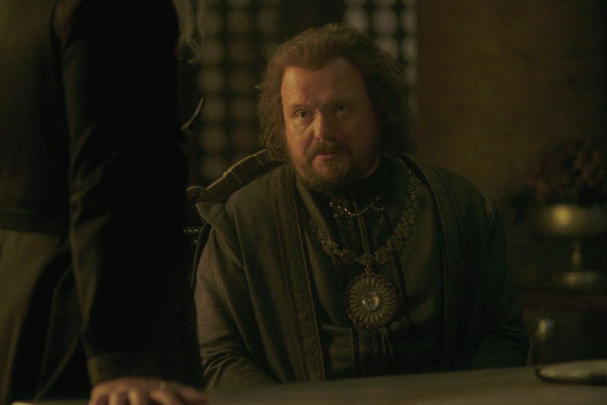 Lord Lyonel Strong sits on a table opposite Viserys, whose right arm is seen on the left of the image, in episode 2 of House of the Dragon.